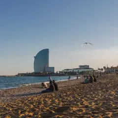 Ludzie odpoczywają na plaży w Barcelonie, a w tle widać charakterystyczny budynek hotelu i błękitne morze.