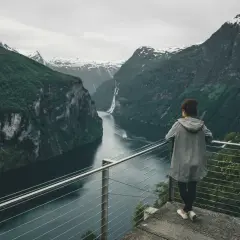 Kobieta podziwia majestatyczne fiordy norweskie, z wodospadem w oddali i ośnieżonymi szczytami gór.