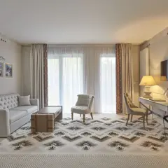 Elegancki pokój w hotelu 5 gwiazdkowym z sofą, fotelem, biurkiem i telewizorem. Wzory na dywanie i zasłonach dodają charakteru.