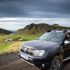 Tanie wycieczki Islandia: Dacia Duster na tle zielonej doliny z farmą i górami. Idealne auto na islandzką przygodę.