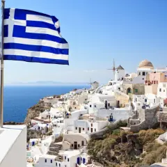 Grecka flaga powiewa nad białymi domkami na Santorini. Idealne miejsce, by zastanowić się, co zabrać na wakacje do Grecji all inclusive.