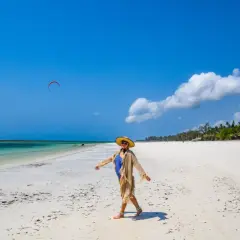 Kobieta w stroju kąpielowym i kapeluszu cieszy się słońcem na piaszczystej plaży Diani Beach. Na niebie czerwony latawiec do kitesurfingu.
