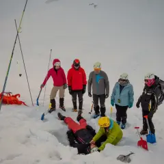 Grupa ćwiczy abc lawinowe. Jedna osoba leży w śniegu, inni ją odkopują. W tle widać innych narciarzy.