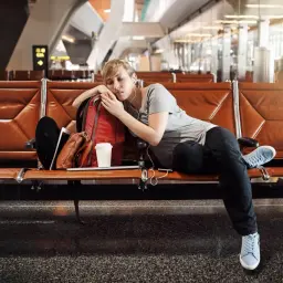 Zmęczona podróżniczka śpi na lotnisku, próbując pokonać jetlag. Obok niej plecak i kubek kawy.