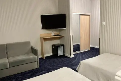 Pokój w sanatorium Arka Kołobrzeg: dwa łóżka, sofa, szafa z lustrem, telewizor i mini lodówka.