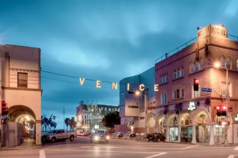 Napis "VENICE" rozświetla nocne niebo nad ulicą w Venice Beach. Widać budynki, samochody i znaki drogowe.