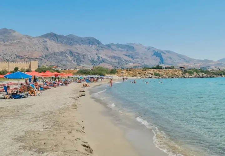 Słoneczna plaża w Frangokastello z turkusową wodą, leżakami i widokiem na zabytkowy zamek.