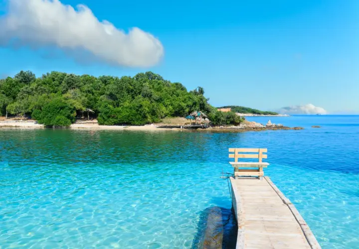 Albania - wczasy samolotem all inclusive. Drewniane molo z ławką prowadzi do krystalicznie czystej wody. Zielona wyspa w tle.