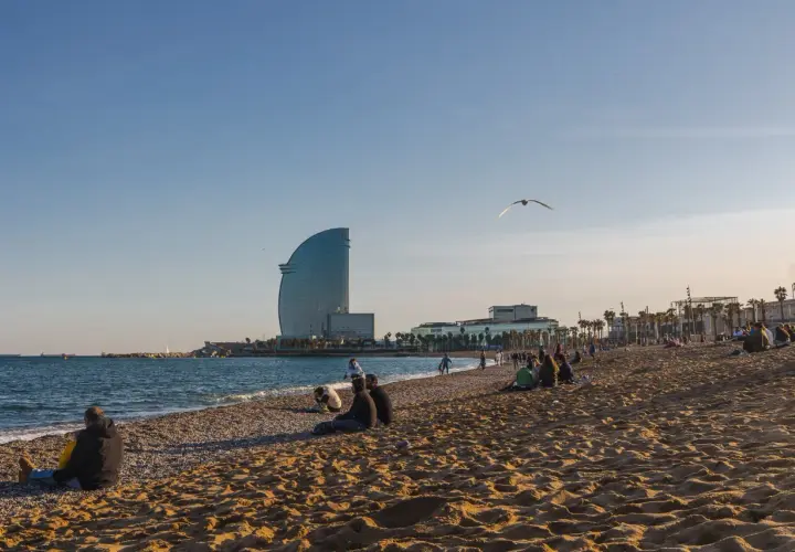 Ludzie odpoczywają na plaży w Barcelonie, a w tle widać charakterystyczny budynek hotelu i błękitne morze.