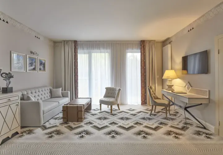 Elegancki pokój w hotelu 5 gwiazdkowym z sofą, fotelem, biurkiem i telewizorem. Wzory na dywanie i zasłonach dodają charakteru.