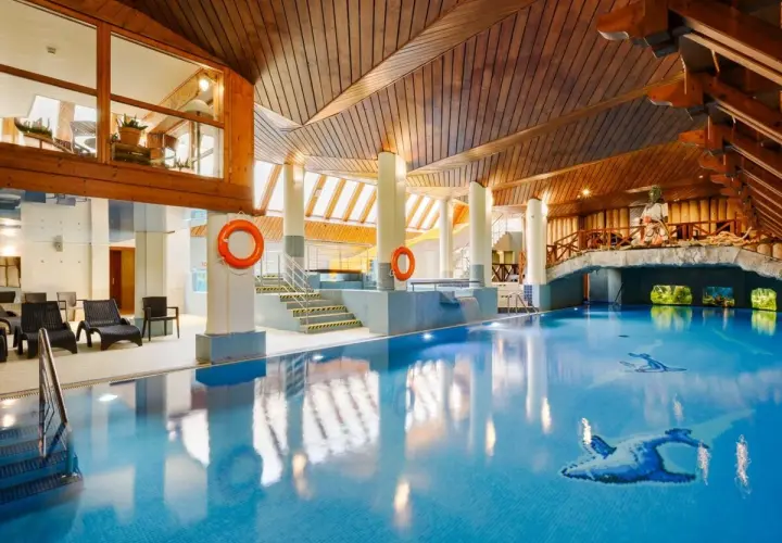 Basen w hotelu Belvedere Resort & Spa z drewnianym sufitem i mostkiem. Woda jest błękitna, a na dnie widać mozaiki z ptakami.