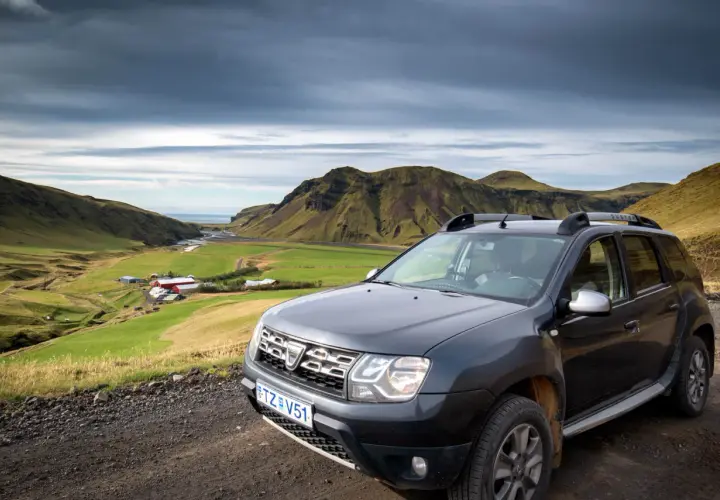 Tanie wycieczki Islandia: Dacia Duster na tle zielonej doliny z farmą i górami. Idealne auto na islandzką przygodę.