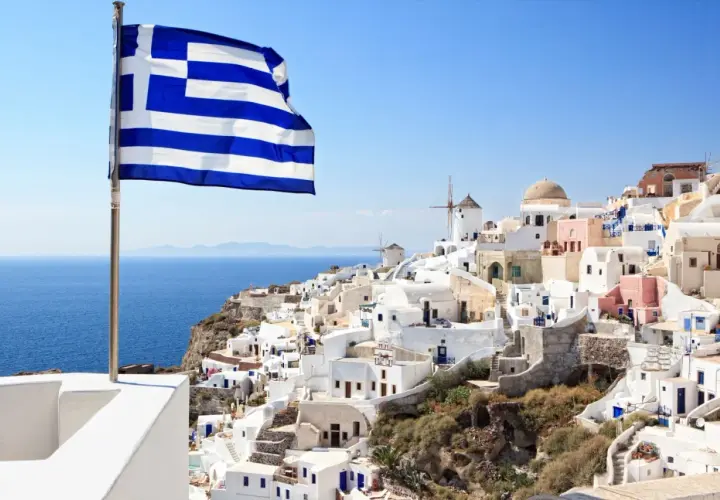 Grecka flaga powiewa nad białymi domkami na Santorini. Idealne miejsce, by zastanowić się, co zabrać na wakacje do Grecji all inclusive.