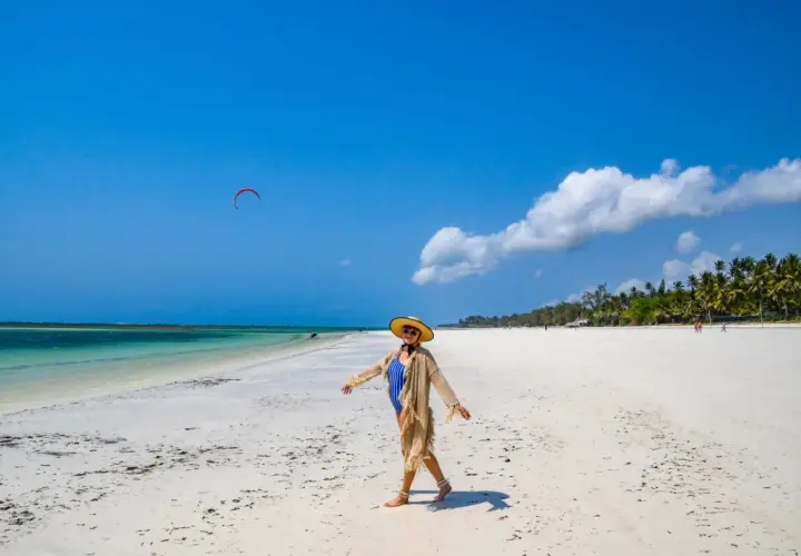 Kobieta w stroju kąpielowym i kapeluszu cieszy się słońcem na piaszczystej plaży Diani Beach. Na niebie czerwony latawiec do kitesurfingu.