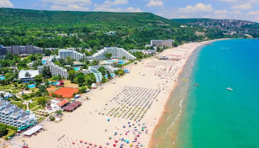 Słoneczne bułgarskie plaże tętnią życiem. Widać hotele, baseny, korty tenisowe i mnóstwo kolorowych parasoli na piaszczystym brzegu.