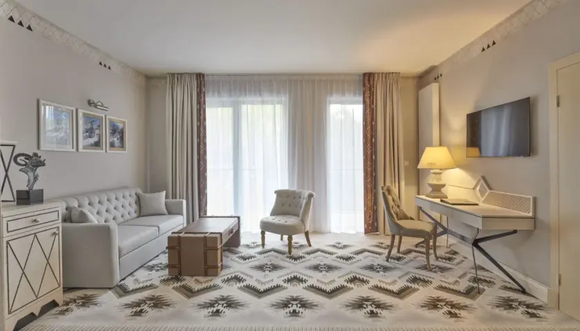 Elegancki pokój hotelowy z sofą, fotelem, biurkiem i telewizorem. Wnętrze w stylu 4-gwiazdkowym, z dywanem o geometrycznym wzorze.