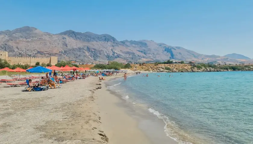 Słoneczna plaża w Frangokastello z turkusową wodą, leżakami i widokiem na zabytkowy zamek.