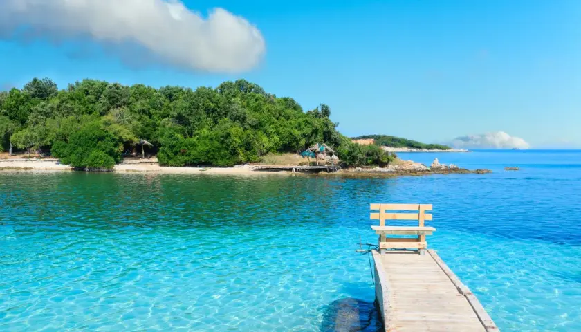 Albania - wczasy samolotem all inclusive. Drewniane molo z ławką prowadzi do krystalicznie czystej wody. Zielona wyspa w tle.