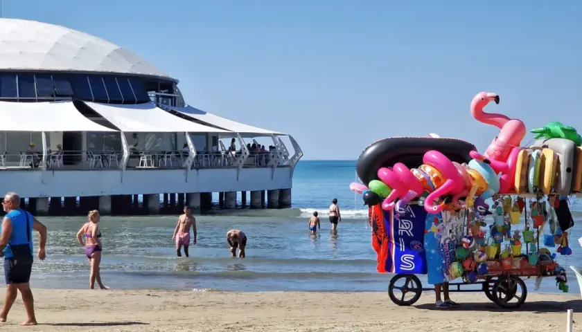 Słoneczna durres plaża z wózkiem pełnym dmuchanych zabawek i restauracją na wodzie.