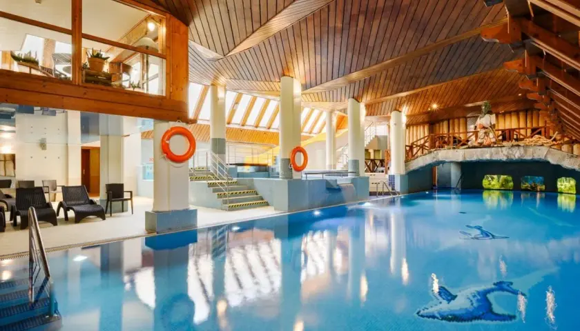 Basen w hotelu Belvedere Resort & Spa z drewnianym sufitem i mostkiem. Woda jest błękitna, a na dnie widać mozaiki z ptakami.