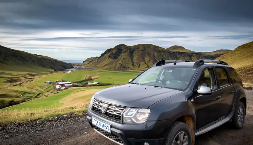 Tanie wycieczki Islandia: Dacia Duster na tle zielonej doliny z farmą i górami. Idealne auto na islandzką przygodę.