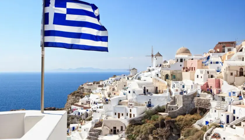 Grecka flaga powiewa nad białymi domkami na Santorini. Idealne miejsce, by zastanowić się, co zabrać na wakacje do Grecji all inclusive.