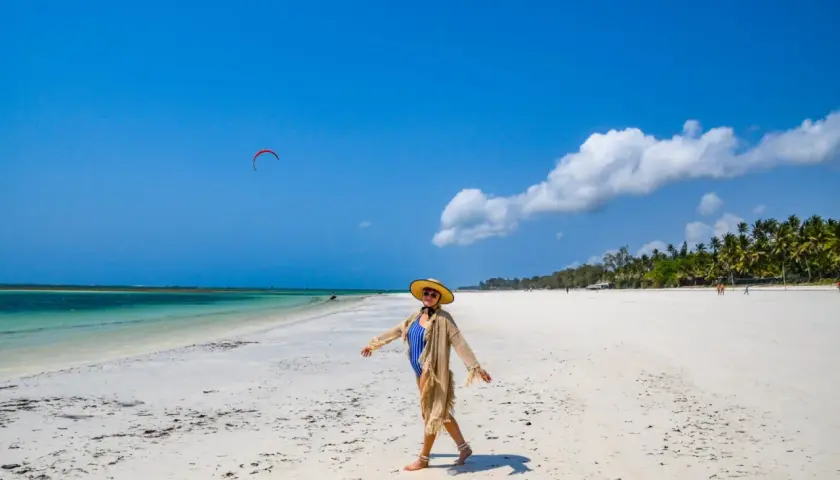 Kobieta w stroju kąpielowym i kapeluszu cieszy się słońcem na piaszczystej plaży Diani Beach. Na niebie czerwony latawiec do kitesurfingu.