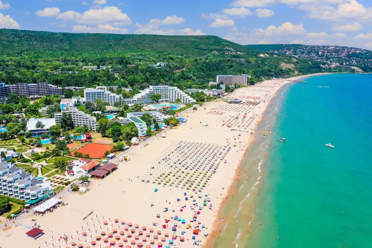 Słoneczne bułgarskie plaże tętnią życiem. Widać hotele, baseny, korty tenisowe i mnóstwo kolorowych parasoli na piaszczystym brzegu.