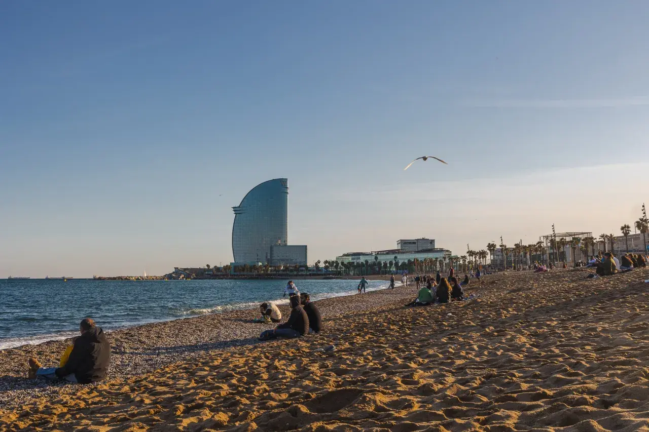Ludzie odpoczywają na plaży w Barcelonie, a w tle widać charakterystyczny budynek hotelu i błękitne morze.