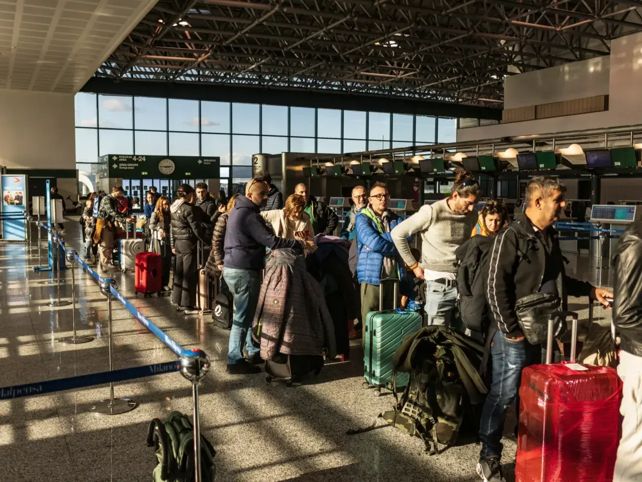 Kolejka pasażerów z bagażami na lotnisku we Włoszech. W tle widać stanowiska odprawy.