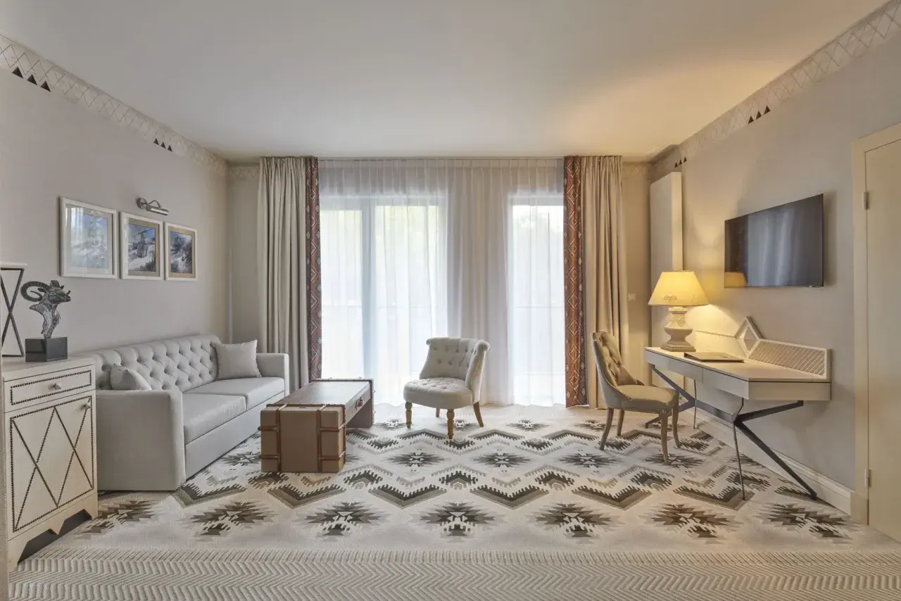 Elegancki pokój w hotelu 5 gwiazdkowym z sofą, fotelem, biurkiem i telewizorem. Wzory na dywanie i zasłonach dodają charakteru.