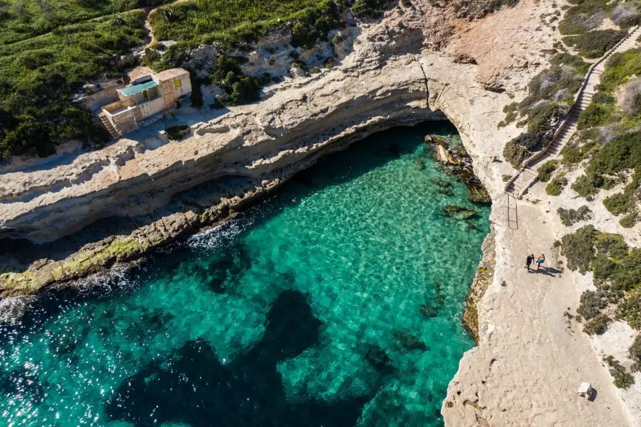 Malta plaże: turkusowa woda, skaliste wybrzeże i mały domek na klifie. Dwie osoby spacerują ścieżką.