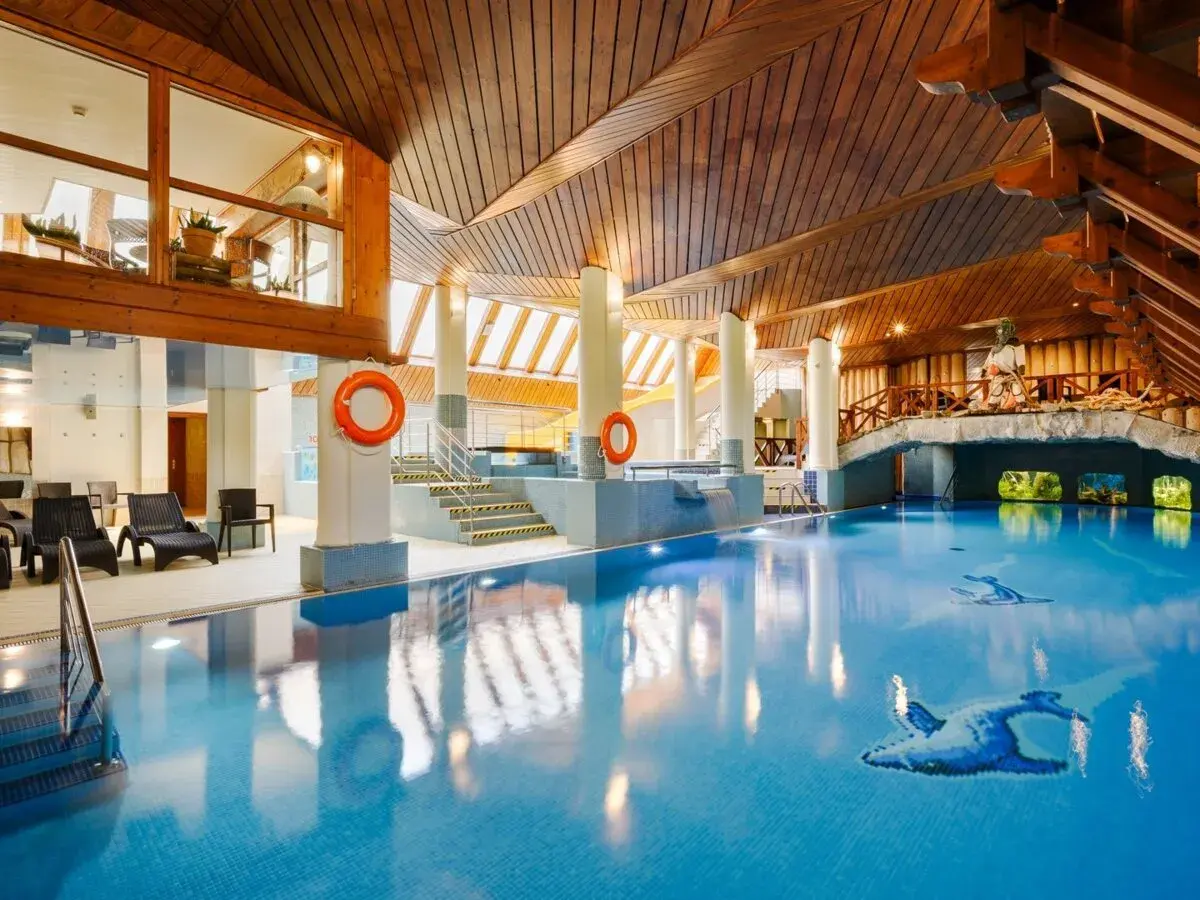 Basen w hotelu Belvedere Resort & Spa z drewnianym sufitem i mostkiem. Woda jest błękitna, a na dnie widać mozaiki z ptakami.