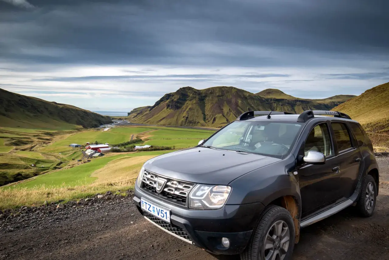 Tanie wycieczki Islandia: Dacia Duster na tle zielonej doliny z farmą i górami. Idealne auto na islandzką przygodę.