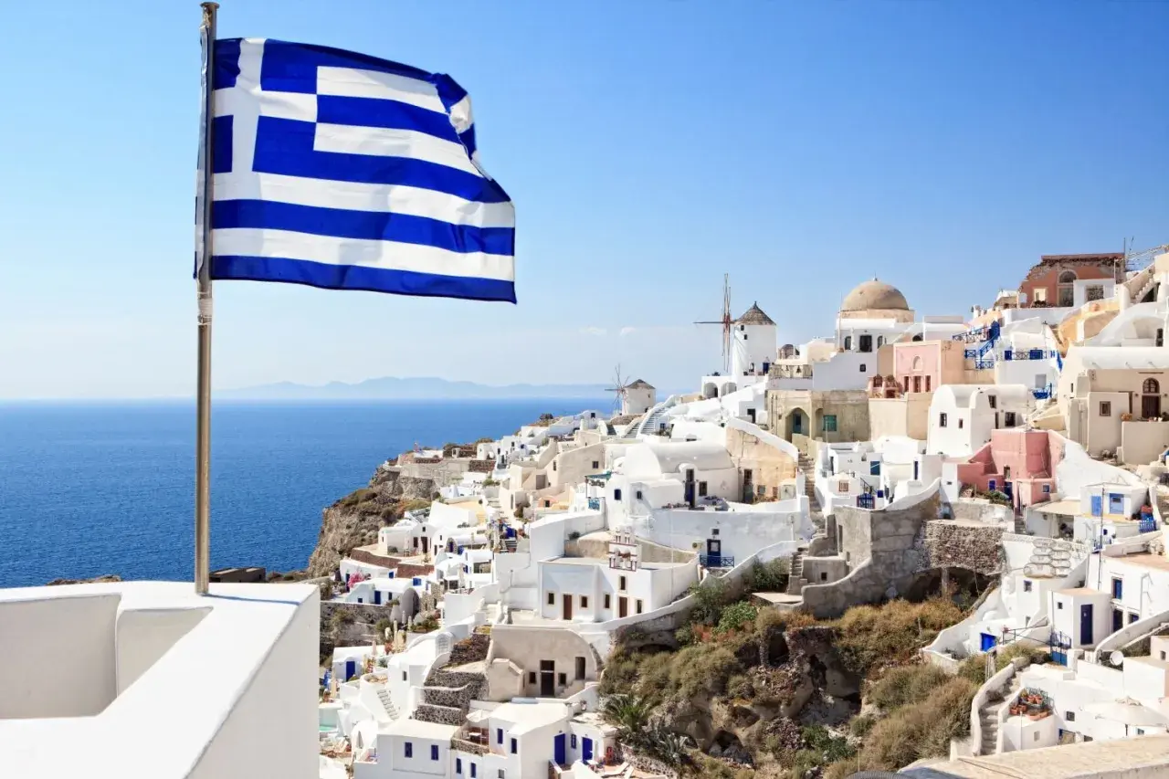 Grecka flaga powiewa nad białymi domkami na Santorini. Idealne miejsce, by zastanowić się, co zabrać na wakacje do Grecji all inclusive.