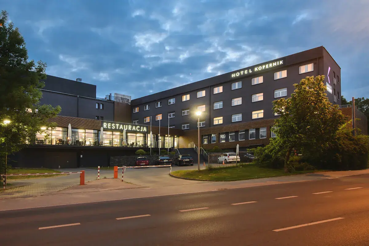 Hotel Kopernik Olsztyn z neonowym napisem "RESTAURACJA" i oświetlonymi oknami. Zapraszamy na nocleg i pyszne jedzenie.