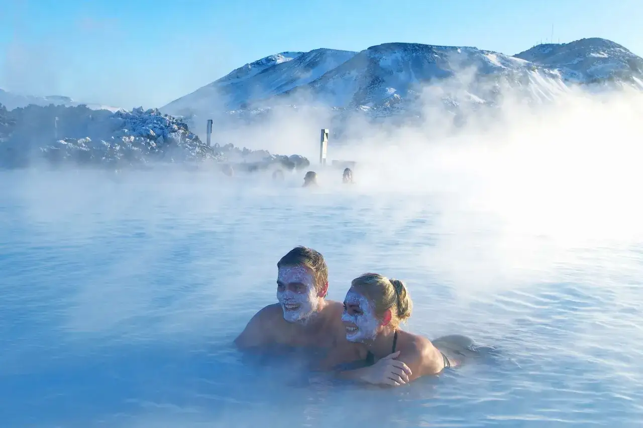 Para relaksuje się w Blue Lagoon, z maskami na twarzach, w otoczeniu pary i ośnieżonych gór.