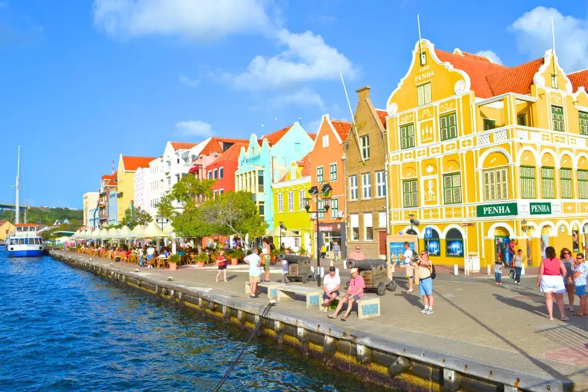 Kolorowe kamienice nad wodą, idealne na wakacje na Curacao. Ludzie spacerują, siedzą przy stolikach, ciesząc się słońcem.