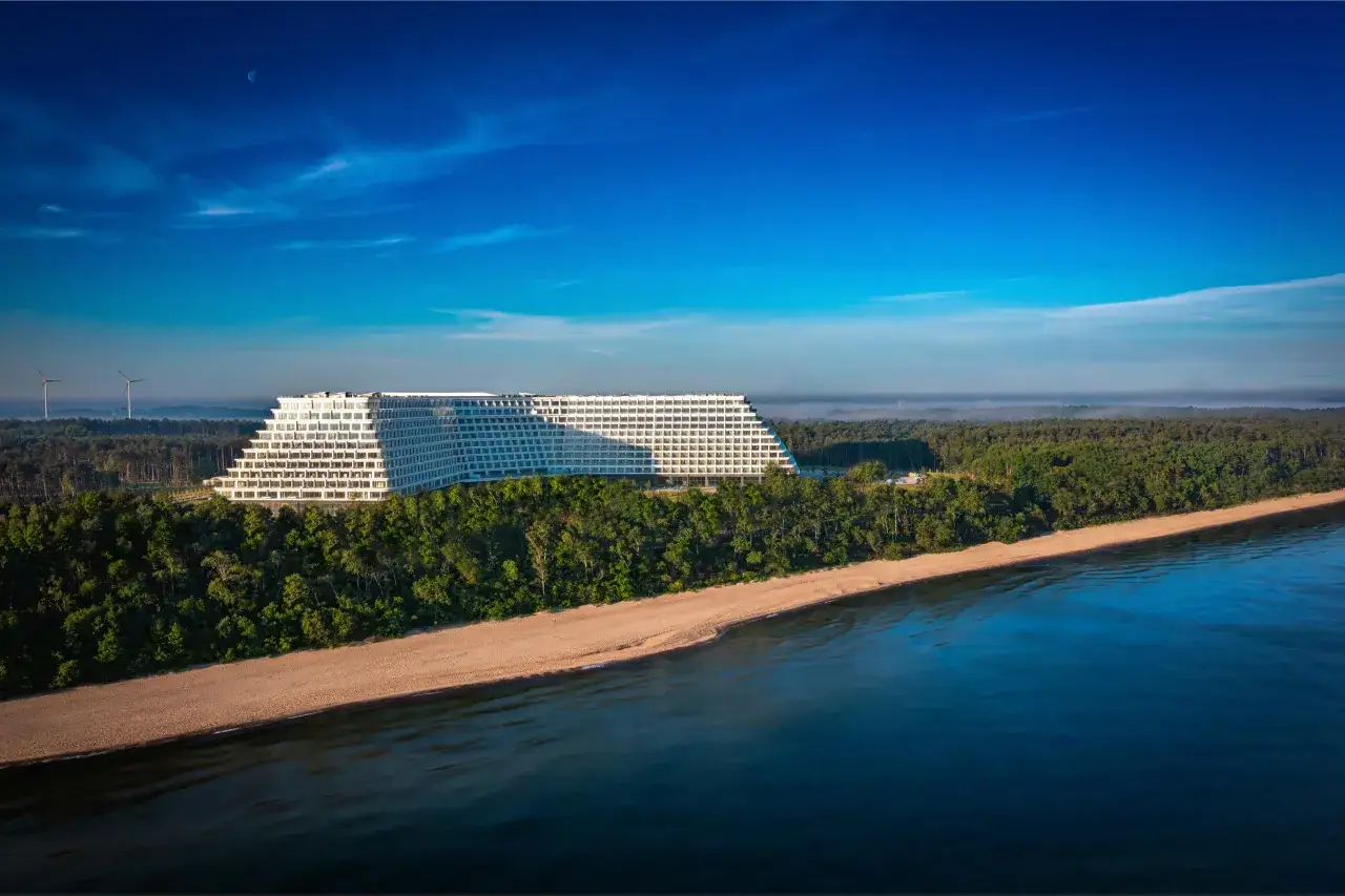 Gołębiewski nad morzem, z widokiem na piaszczystą plażę i las. W oddali widać turbiny wiatrowe i księżyc na błękitnym niebie.