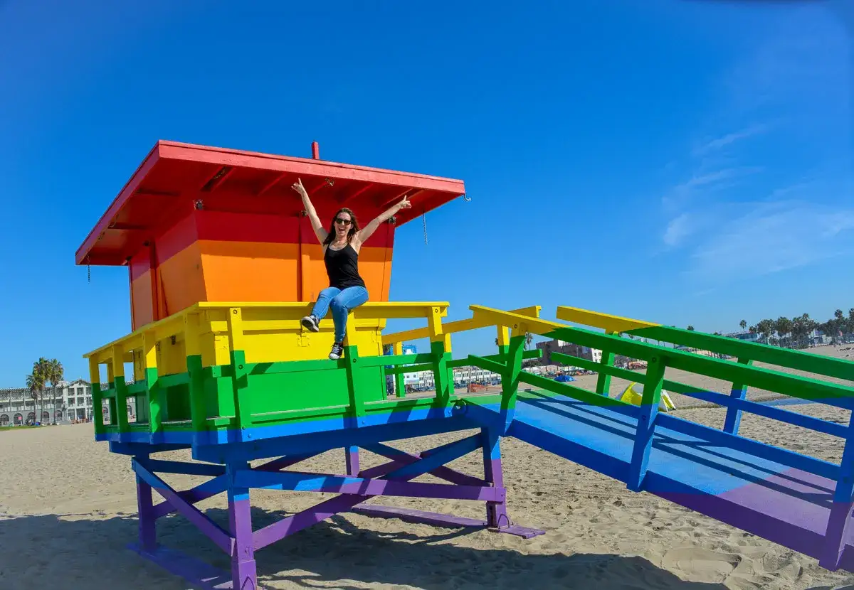 Radosna kobieta siedzi na tęczowej wieży ratownika na Venice Beach.