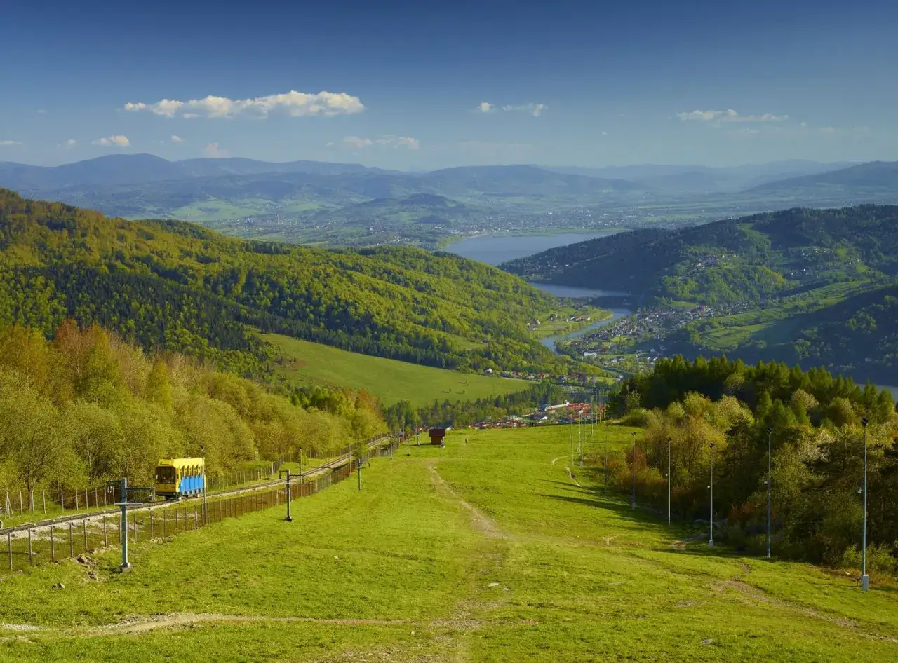 Widok na zielone Beskidy, jezioro i miasteczko. Kolejka wjeżdża na szczyty, a w oddali widać kolejne pasma górskie.