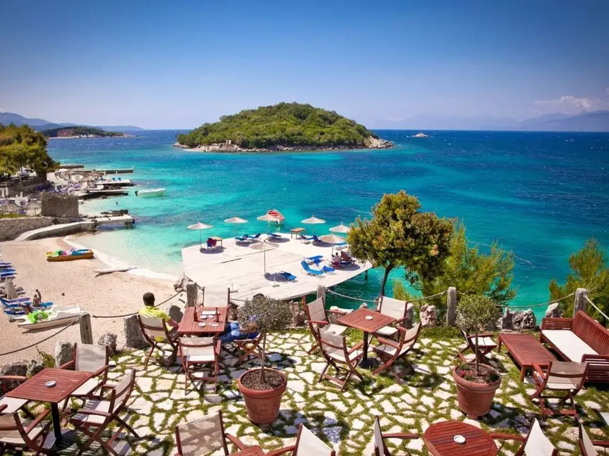 Albania - wczasy samolotem all inclusive. Widok na turkusowe morze, plażę z leżakami i kawiarnię z widokiem na wyspę. Idealne miejsce na relaks.
