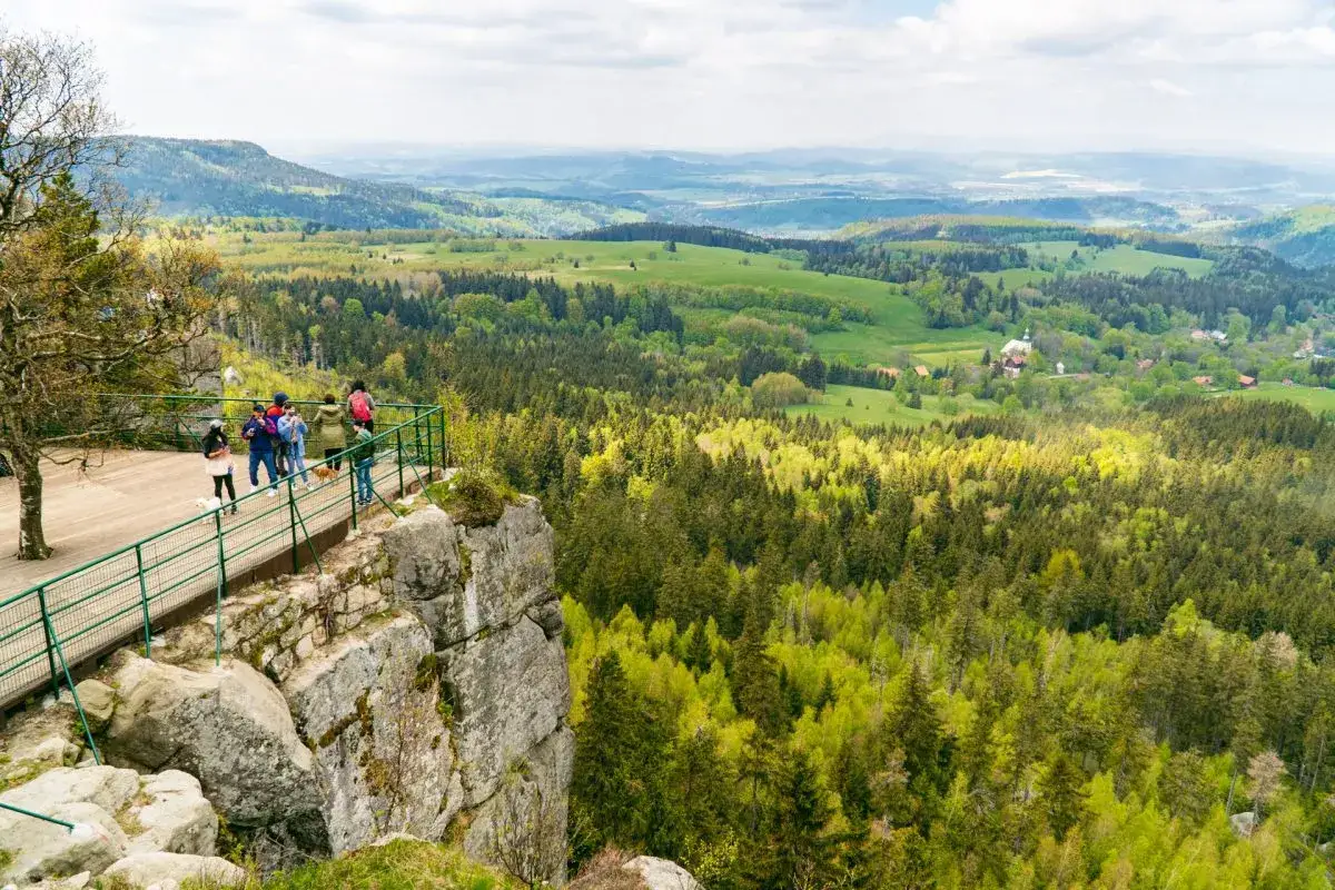 Widok z platformy na skalnym urwisku na zielone wzgórza i lasy Kotliny Kłodzkiej. Grupa turystów podziwia panoramę.