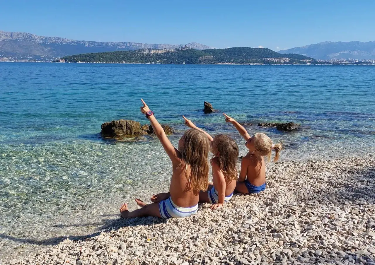 Trzy dziewczynki siedzą na kamienistej plaży w Chorwacji, wskazując palcami na morze. Zastanawiają się, ile kosztują wakacje w Chorwacji dla 4 osób.