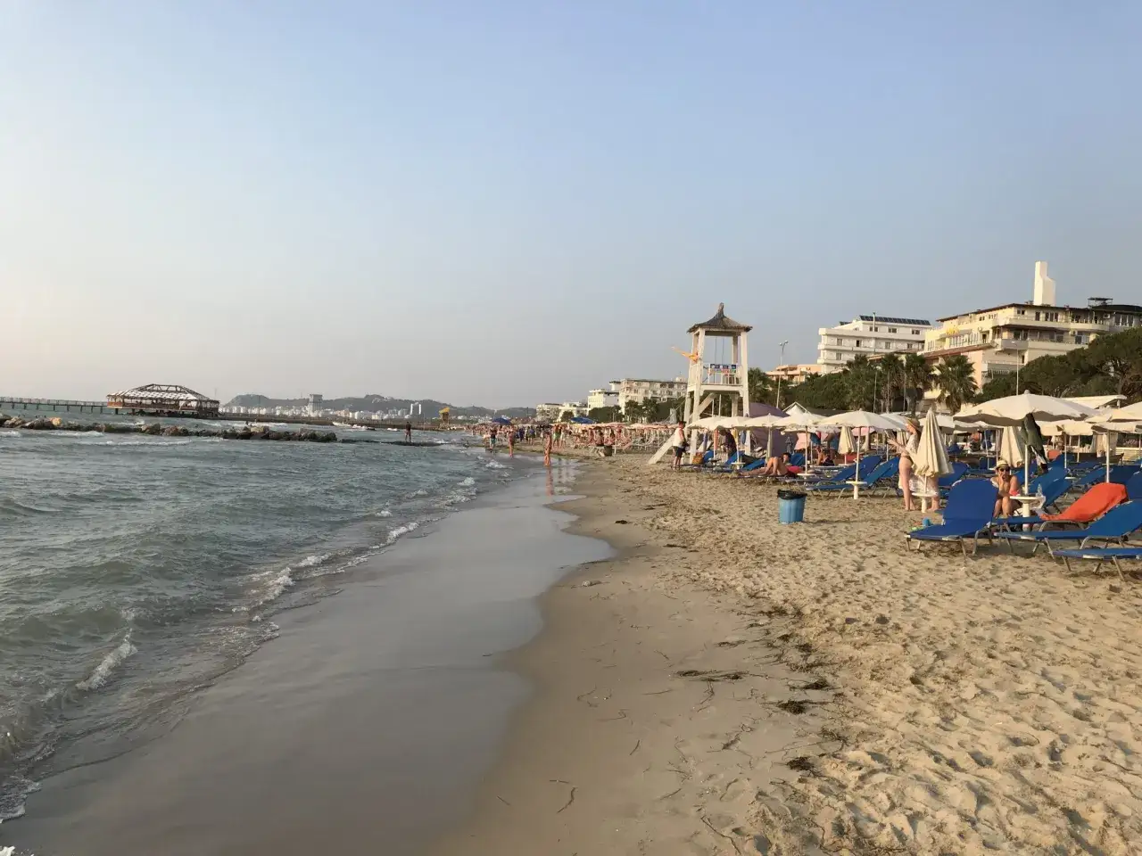 Słoneczna durres plaża z leżakami, parasolami i budką ratownika. W tle widać morze i budynki.