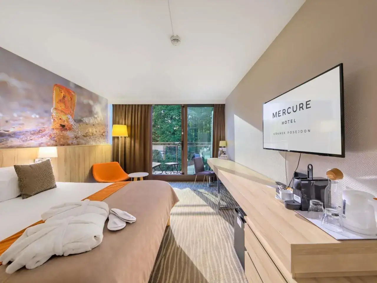 Pokój w hotelu Posejdon Gdańsk z łóżkiem, szlafrokiem i kapciami. Na ścianie mural z bursztynem, a naprzeciwko telewizor z logo Mercure.