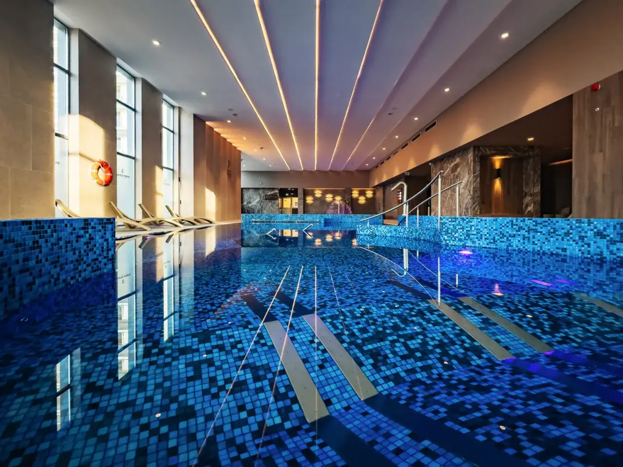 Nowoczesny hotel nad morzem z basenem krytym. Błękitne płytki, leżaki i światło tworzą relaksującą atmosferę.