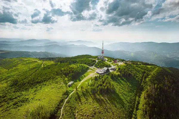 Widok z lotu ptaka na zielone, zalesione **beskidy szczyty** z wieżą telekomunikacyjną i budynkami na jednym ze wzniesień.